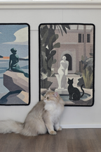 Cat sitting on a wooden floor with two wall hangings featuring artistic depictions of people and a black cat.