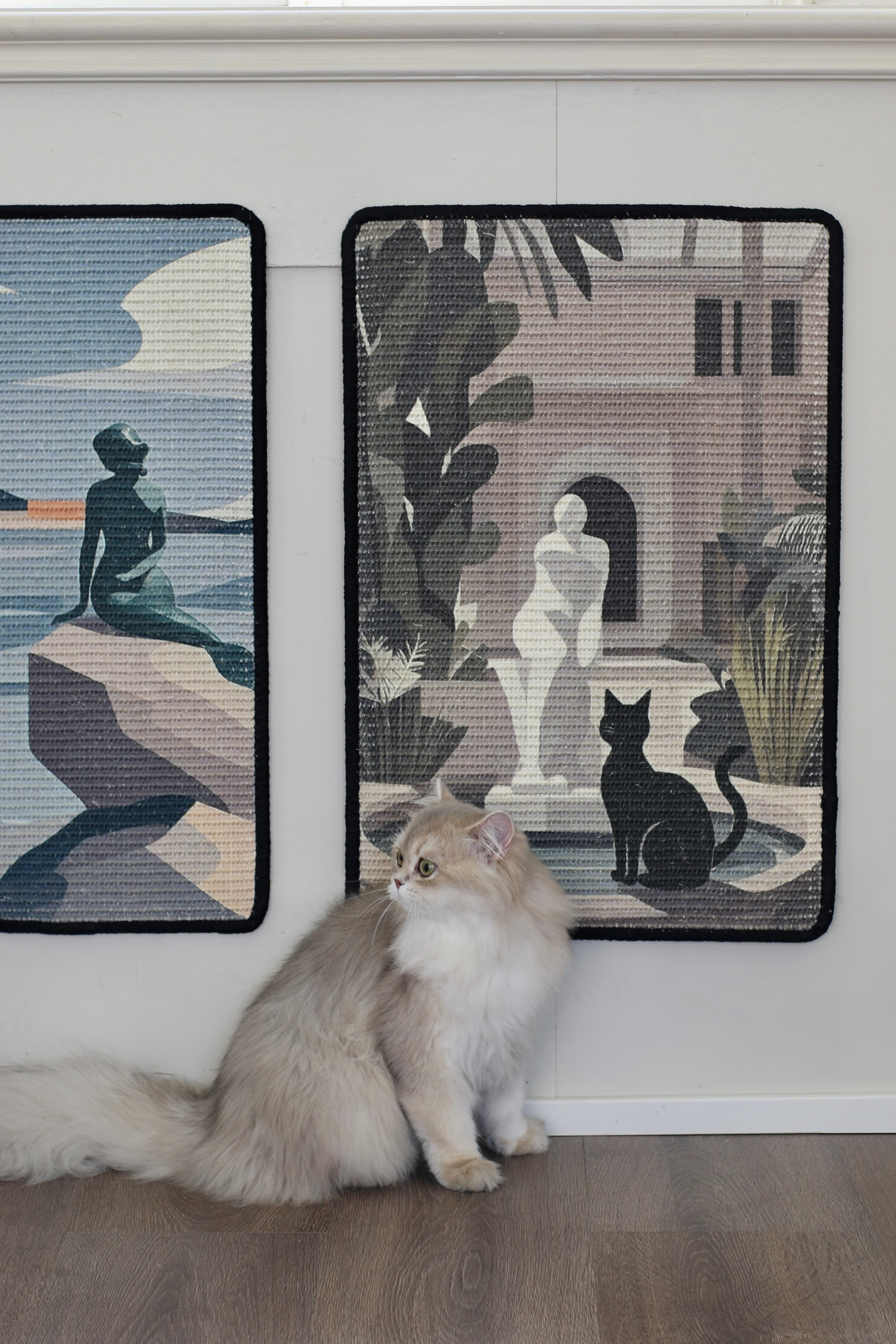 Cat sitting on a wooden floor with two wall hangings featuring artistic depictions of people and a black cat.